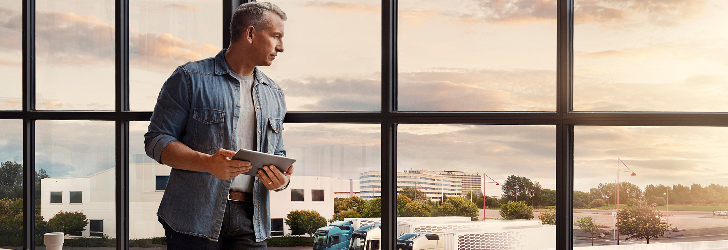 Muž držiaci tablet stojí pri okne a pozerá sa dole na svoj park nákladných vozidiel