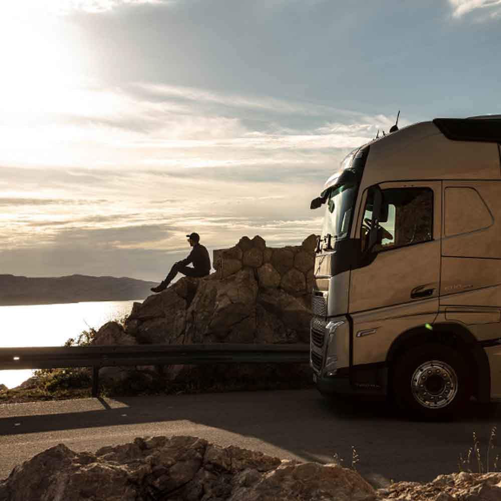 Vodič sedí pri skale s výhľadom na more pri západe slnka a jeho Volvo FH je zaparkované vedľa neho